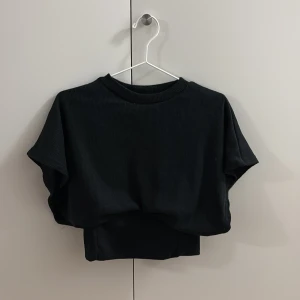 Svart croppad topp från Missguided - Säljer en svart croppad topp från Missguided med ribbad struktur. Toppen har rund halsringning och korta ärmar, perfekt för en chill och stilren look. Materialet är mjukt och stretchigt, vilket gör den skön att bära. Passar dig som gillar minimalistisk stil.