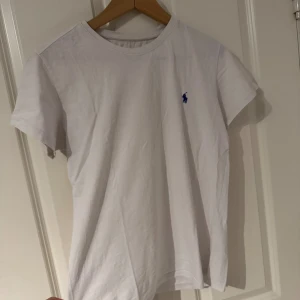 Vit t-shirt från Polo Ralph Lauren - Vit klassisk t-shirt från Polo Ralph Lauren i mjuk bomull. T-shirten har korta ärmar och en rund halsringning. På bröstet finns den ikoniska blå Polo-loggan broderad. Perfekt basic-plagg med snygg passform.