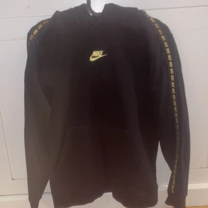 Svart Nike hoodie med gula detaljer - Svart hoodie från Nike med gul logga på bröstet och gul Nike-text längs ärmarna. Klassisk känguruficka framtill och huva. Perfekt för en sportig och avslappnad stil. Tillverkad i mjuk bomullsmix för extra komfort.