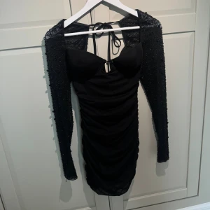 Svart kort klänning från Oh Polly - Supersnygg svart kort klänning från Oh Polly med långa ärmar täckta av små pärlor och paljetter. Klänningen har snörning i ryggen, figurnära passform och rynkade detaljer längs hela kroppen. Perfekt för dig som vill sticka ut.