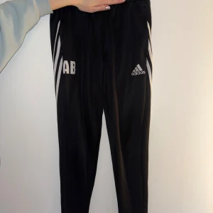 Svarta Adidas träningsbyxor med dragkedja - Svarta träningsbyxor från Adidas med vita ränder längs sidorna och broderad logga. Byxorna har smal passform, dragkedja vid benslut och är tillverkade i lätt polyester med Climalite-teknologi. Perfekta för sport eller chill.