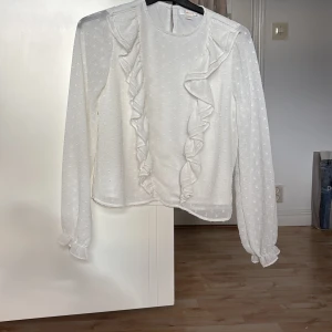 Vit volangblus från Gina Tricot - Supersöt vit blus från Gina Tricot med volangdetaljer framtill och diskret prickigt mönster i tyget. Blusen har långa ärmar med resår och liten volang vid ärmslut. Perfekt för dig som gillar romantisk och trendig stil.