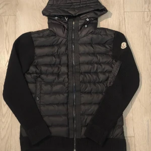 Svart Moncler Cardigan M - Svart Cardigan från Moncler storlek Mmed stickade ärmar och vadderad kropp. Jackan har luva med dragkedja framtill och fickor med knappstängning. Moncler-logga på vänster ärm och extra dragkedjeficka på ärmen. Nfc tagg, lappar och certifikat. Oanvänd i perfekt skick! Meddela för frågor!