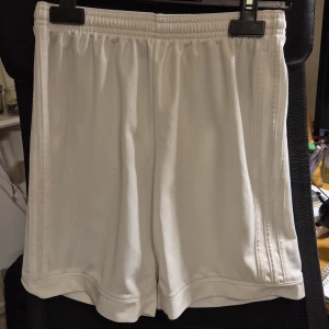 Vita Adidas shorts Climalite 13-14Y/L - Vita sportiga shorts från Adidas med elastisk midja och diskreta ränder längs sidorna. Tillverkade i lätt och svalt Climalite-polyestertyg, perfekta för träning. Adidas-logga baktill och klassisk stil. Liten svart fläck, annars väldigt fint skick.