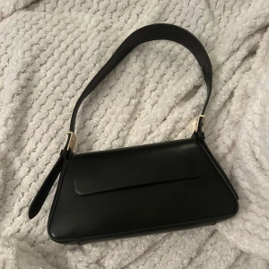 Svart axelväska i skinn med gulddetaljer - Stilren svart axelväska i skinn med en geometrisk form och guldfärgade metalldetaljer vid axelremmen. Väskan har ett lock med magnetstängning och ett litet fack inuti. 