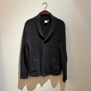 Mörkgrå stickad kofta från Boomerang - Säljer en mörkgrå stickad kofta från Boomerang med snygg kabelstickning, bred krage och knappar framtill. Koftan har två fickor och mockadetaljer på armbågarna för en extra touch. Perfekt för lager-på-lager under hösten och vintern. Hör av er vid frågor eller fler bilder