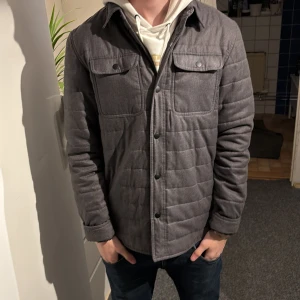 Grå quiltad overshirt med knappar - Snygg grå overshirt med quiltad design och tryckknappar framtill. Jackan har två bröstfickor med lock och lång ärm. Perfekt lager-på-lager-plagg med stilren look och normal passform.