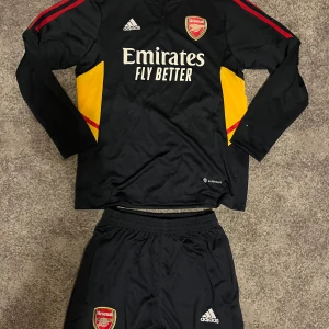Arsenal långärmad träningsset Adidas - Bara tröjan: 249kr Bara byxorna: 199kr Set: 400kr       Snygg Arsenal långärmad träningströja från Adidas med klubbmärke och Emirates-tryck på bröstet. Svart med röda detaljer på ärmarna och gula paneler på sidorna. Tillverkad i funktionsmaterialet Aeroready som andas. Perfekt för fotbollsträning eller supporterliv.