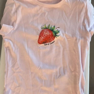 Ljusrosa t-shirt med jordgubbstryck - Söt ljusrosa t-shirt med ett färgglatt jordgubbstryck och texten 'Taste of Summer' på bröstet. Klassisk rund halsringning och korta ärmar. Perfekt för dig som gillar somriga och lekfulla plagg.