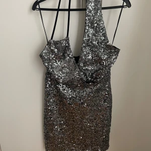 Glittrig silver ”one shoulder” klänning - Säljer en glittrig silverfärgad kortklänning med ”one shoulder” och figurnära passform. Klänningen är täckt av paljetter och har en djup urringning samt tunna axelband på ena sidan. Perfekt för dig som vill sticka ut och glänsa på festen. Den är endast använd 1 gång så den är i ett fint skick, säljer då den inte kommer till användning. 💕hör av er vid frågor 