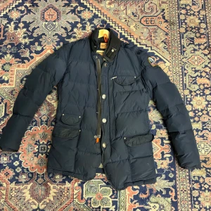 Mörkblå dunjacka från Parajumpers - Snygg mörkblå dunjacka från Parajumpers med flera fickor, silverfärgade knappar och dragkedja. Jackan har hög krage med orange detalj och patch på ärmen. Perfekt för kalla dagar och har en modern, lite längre passform.