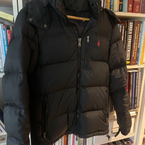 Svart pufferjacka från Polo Ralph Lauren - Svart pufferjacka från Polo Ralph Lauren med huva och tvåvägsdragkedja framtill. Jackan har broderad röd logga på bröstet, flera fickor med dragkedja och quiltad design. Perfekt för kalla dagar och har en clean, klassisk look. Mycket bra skick förutom små defekter på baksida armar. Kan fixas enkelt med patches, skriv för info angående det. Pris kan diskuteras.  