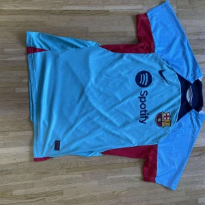 Barcelona Nike fotbollströja ljusblå - Barcelona fotbollströja från Nike i ljusblå med röda detaljer på ärmarna och klubbmärke på bröstet. Tröjan har korta ärmar, rund hals och tryck med Spotify-logga. Materialet är lätt och andas, perfekt för match eller träning.