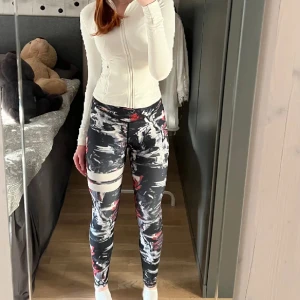 Stronger träningstights och topp - Stronger träningstights och matchande cropped topp med abstrakt mönster i svart, vitt, rött och grått. Hög midja och stretchigt material som passar perfekt till träning. Logga på linningen och cool design som sticker ut.