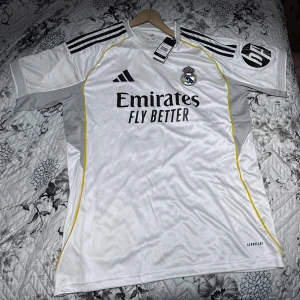 Real Madrid  - Splitterny Real Madrid matchtröja från Adidas i vitt med svarta detaljer och gula linjer. Tryck med 'Emirates Fly Better' på bröstet, klubbmärke och nummer 12 med namnet Trent på ryggen. Kortärmad modell i lätt och ventilerande material.