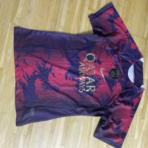 PSG x Nike röd-blå fotbollströja - Säljer en Paris Saint-Germain fotbollströja från Nike med Qatar Airways-tryck. Tröjan har ett unikt abstrakt mönster i mörkblått och rött, korta ärmar och rund hals. Materialet är lätt och andas, perfekt för träning eller match. PSG-logga och Nike Swoosh på bröstet.