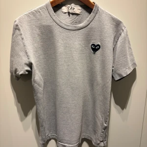 Grå Comme des Garçons Play t-shirt - Grå t-shirt från Comme des Garçons Play med klassisk svart hjärta med ögon broderat på bröstet. T-shirten har rund halsringning och korta ärmar. Tillverkad i mjuk bomull för en skön och avslappnad känsla.