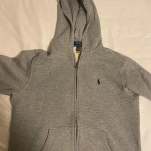 Grå hoodie från Polo Ralph Lauren - Klassisk grå hoodie från Polo Ralph Lauren med dragkedja framtill, huva och kängurufickor. Broderad marinblå logga på bröstet. Mjuk insida och långärmad modell, perfekt för chill dagar.