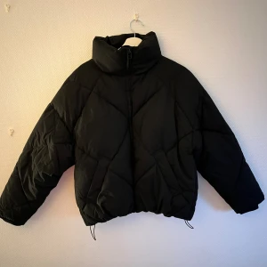Svart pufferjacka från Stay, S - Svart pufferjacka från Stay i storlek S. Jackan har hög krage, dragkedja framtill och quiltad design med snygga sömmar. Modellen är lite oversized och har dragsko nertill för att justera passformen. Perfekt för kalla dagar.