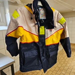 Färgglad vindjacka med reflexdetaljer - Cool vindjacka i färgerna gul, marinblå och beige med röda detaljer. Jackan har huva, stora fickor och flera reflexdetaljer på axlar och huva. Perfekt för dig som vill sticka ut och synas. Materialet är syntet och jackan har dragkedja framtill.