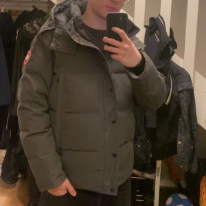 Mörkgrå Canada Goose jacka - Säljer en mörkgrå jacka från Canada Goose med huva och klassisk logopatch på ärmen. Jackan har quiltad design, dragkedja och knappar framtill samt två stora fickor. Perfekt för kalla vinterdagar och riktigt snygg till streetwear. Ett problem är att dom 2 knapparna längst ner har gått sönder och har inte päls därför släjs den billigt