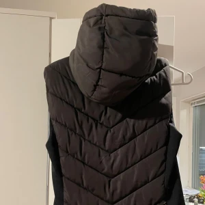 Svart väst med luva  - Snygg svart dunväst med huva och dragkedja framtill. Västen har ett quiltat mönster i sicksack och är perfekt för lager-på-lager. Passar dig som gillar en stilren look och vill hålla värmen utan att tumma på stilen.