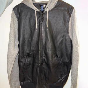 Svart och grå jacka H&M storlek M - Snygg tunn jacka från H&M i storlek M med svart perforerat skinnliknande material på kroppen och gråa bomullsärmar samt huva. Jackan har dragkedja framtill, snörning vid huvan och två fickor med dragkedja. Perfekt för dig som gillar streetstyle.