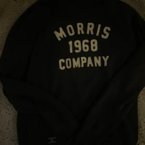 Svart stickad tröja Morris Gents S - Svart stickad tröja från Morris Gents med vit text 'Morris 1968 Company' framtill. Klassisk rund halsringning och långa ärmar. Perfekt för dig som gillar stilren och sportig look.