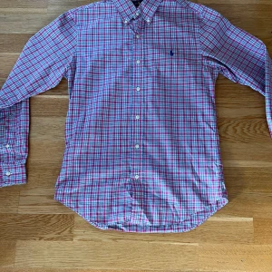 Rutig skjorta från Ralph Lauren - Rutig skjorta från Ralph Lauren i slim fit med klassisk krage och knappar framtill. Skjortan har ett mönster i rött, blått och vitt samt broderad logga på bröstet. Tillverkad i mjuk bomull och har långa ärmar med manschettknappar.