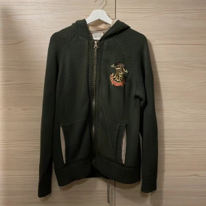 Mörkgrön hoodie från Polo Ralph Lauren - Mörkgrön hoodie från Polo Ralph Lauren med broderad logga på bröstet. Tröjan har dragkedja, två fickor fram och ribbade muddar. Tillverkad i mjuk bomull med en touch av kashmir för extra komfort. Perfekt för höst och vinter.