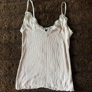 Ljusbeige spetslinnetopp från Amisu - Supersöt ljusbeige linnetopp från Amisu med tunna axelband och v-ringning dekorerad med spets. Ribbad struktur i tyget och fina volangdetaljer längst ner. Perfekt för sommardagar eller att styla lager på lager.