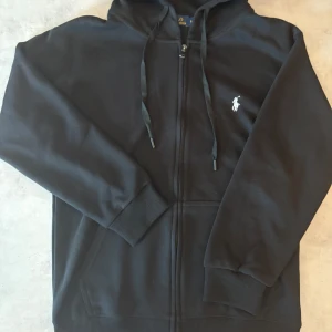 Svart hoodie från Polo Ralph Lauren - Svart hoodie från Polo Ralph Lauren med dragkedja, huva med snörning och klassisk vit logga på bröstet. Tröjan har ribbade muddar och midja samt praktiska fickor framtill. Perfekt för en chill och stilren look.