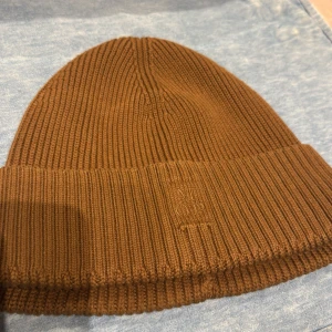 Brun ribbstickad mössa Marc O'Polo - Snygg brun ribbstickad mössa från Marc O'Polo. Tillverkad i en mjuk mix av bomull och kashmir för extra komfort. Klassisk uppvikt kant med diskret broderad logga framtill. Perfekt accessoar för kyliga dagar.