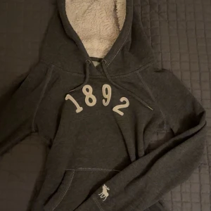 Mörkgrå hoodie Abercrombie & Fitch - RESERVERAD!                                                        Mysig mörkgrå hoodie från Abercrombie & Fitch med broderat 1892 framtill. Fodrad huva med fluffigt teddyfoder, snörning i halsen och klassisk känguruficka. Liten vit älg-logga på ärmen. Perfekt för kyliga dagar.