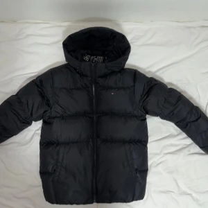 Svart pufferjacka Tommy Hilfiger - Svart pufferjacka från Tommy Hilfiger med huva och dragkedja. Jackan har broderad logga på bröstet och stor logotyp på insidan av kragen. Fodrad och riktigt varm, perfekt för kalla dagar. Klassisk design med två fickor framtill. Storlek S 176cm men den kan passa upp till 185 ganska lätt tycker jag för att jag är 180 och den passar mig ganska bra.