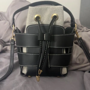 Svart och beige axelväska med snörning - Stilren axelväska i svart skinn med vita kontrastsömmar och detaljer i guld. Väskan har en beige innerpåse i tyg och stängs med snörning. Justerbar axelrem och cool design som sticker ut. 