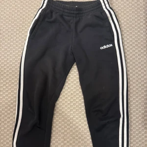 Svarta Adidas mjukisbyxor med ränder - Svarta mjukisbyxor från Adidas med klassiska vita ränder längs sidorna och Adidas-logga på låret. Byxorna har resår i midjan och är tillverkade i mjuk bomullsmix, perfekta för chill eller träning.