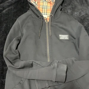 Svart hoodiejacka från Burberry - Svart hoodiejacka från Burberry med dragkedja och snörning i huvan. Insidan har klassiskt rutigt Burberry-mönster i beige, rött och svart. Jackan är tillverkad i bomull och har en stilren logotyp på bröstet. Perfekt för dig som gillar ikoniska detaljer och streetwear. PASSAR L