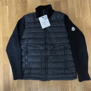 Svart pufferjacka Moncler M - Svart pufferjacka från Moncler med stickade ärmar och quiltad front. Jackan har dragkedja framtill, två sidofickor med dragkedja och Moncler-logga på vänster ärm. Snygg, stilren och perfekt för kyliga dagar.