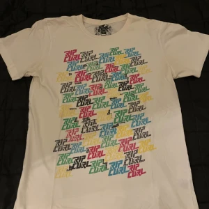 Vit Rip Curl t-shirt med färgglatt tryck - Vit t-shirt från Rip Curl i slim fit-modell. Framsidan har ett stort, färgglatt tryck med Rip Curl-loggan i blått, grönt, gult, svart och rött. Klassisk rund hals och korta ärmar. Perfekt för en avslappnad streetstil.