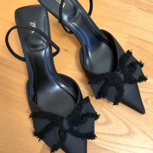 Svarta pumps med rosett från Zara - Snygga svarta pumps från Zara med spetsig tå och dekorativa stora rosetter framtill. Skorna har smal rem runt hälen och låg klack. Tillverkade i ett glansigt tygmaterial som ger en elegant look. Oanvända.