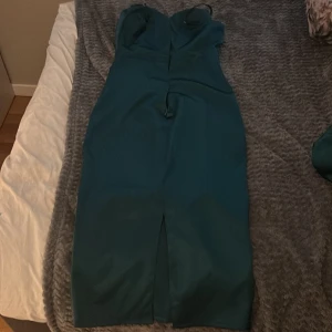 Mörkgrön långklänning med slits - Snygg mörkgrön långklänning med smala axelband och v-ringad omlott upptill. Klänningen har en slits framtill och avtagbara puffärmar som ger en cool touch. Materialet är slätt och har en lätt satinliknande finish som känns lyxig.