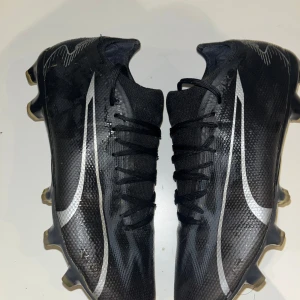 Svarta fotbollsskor från Puma strl 39 - Säljer ett par svarta fotbollsskor från Puma i storlek 39. Skorna har en snygg, sportig design med silvriga detaljer och snörning. Ovandelen är i syntetmaterial och sulan har fasta dobbar för bra grepp på planen. Perfekta för dig som vill ha stilrena och snabba skor på matchen.