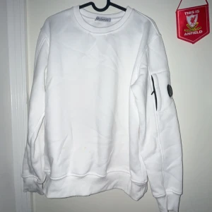 Vit sweatshirt från C.P. Company - Vit sweatshirt från C.P. Company med klassisk rund hals och ribbade muddar. Tröjan har en ikonisk svart linsdetalj på ärmen och en ficka med dragkedja. Tillverkad i mjuk bomullsblandning för en skön och avslappnad look.