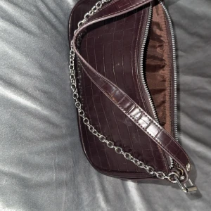 Mörkbrun axelväska med kedjedetalj - Snygg mörkbrun axelväska i croco-mönstrat skinnimitation med silvrig kedja framtill. Väskan har dragkedja upptill och justerbart axelband med metallringar. Perfekt storlek för det viktigaste och ger en trendig vibe till din outfit. De går att ta bort silver kedjan på väskan.