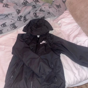 Svart vindjacka från Nike med huva - Svart Nike vindjacka med huva och dragkedja framtill. Jackan har två fickor med dragkedja, ribbade muddar och meshfoder på insidan. Klassisk Nike-logga på bröstet och sportig look. Perfekt för dig som gillar streetwear och enkel stil.