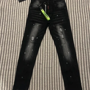 Svarta Dsquared2 jeans med detaljer - Snygga svarta jeans från Dsquared2 med slitningar, vita färgstänk och en grymt cool neongrön nyckelring. Jeansen har smal passform och klassisk femficksdesign. Materialet är bomull med stretch för extra komfort. Perfekta för dig som gillar streetstyle och unika detaljer.