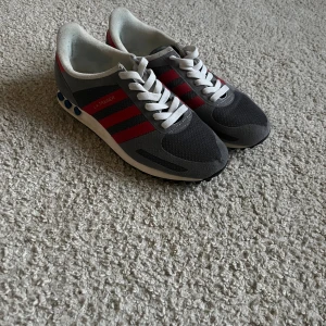 Adidas LA Trainer sneakers grå/röd - Säljer ett par Adidas LA Trainer sneakers i grått och svart med röda ränder. Skorna har vit sula med blå detaljer och klassisk snörning. Ovandelen är i mesh och mocka för en sportig vibe. Perfekta för dig som gillar retrostil och streetwear. Obs, sulan finns inte men det är inget som gör nån skillnad