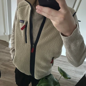 Beige fleeceväst från Patagonia - Säljer en beige fleeceväst från Patagonia med marinblå detaljer och röda dragkedjor. Västen har hög krage, tre fickor med dragkedja och klassisk Patagonia-logga på bröstet. Perfekt för lager-på-lager och chill vardagsstil. Hör av er vid frågor och funderingar😃