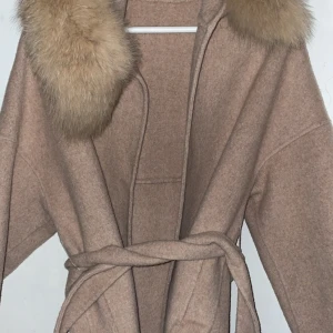 Beige kappa med pälskrage - Säljer en beige kappa med stor fluffig pälskrage och huva. Jackan har knytskärp i midjan, stora fickor framtill och är i en rak, loose passform. Perfekt för dig som vill ha en mysig och snygg look under höst och vinter.